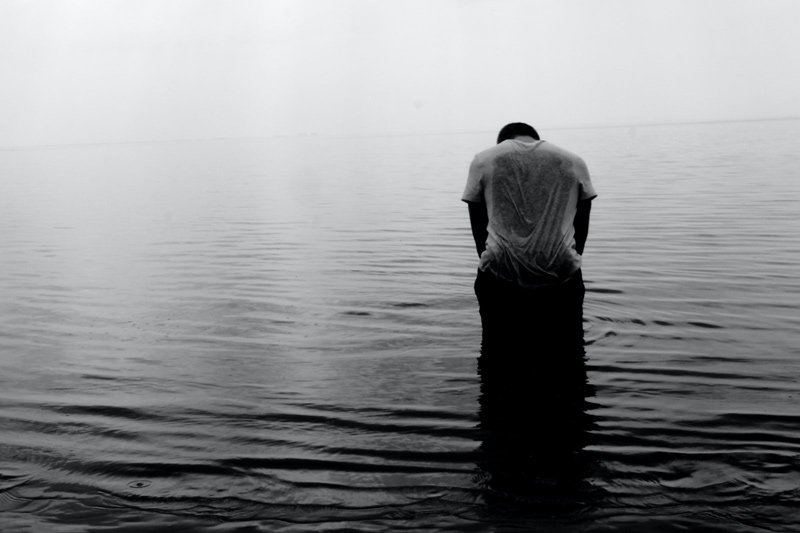 Man standing in a body of water head down facing away from the camera, looking alone and sad