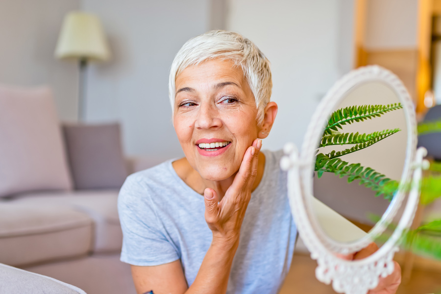 Lady with white hair happily gazes into a mirror whilst holding her cheek